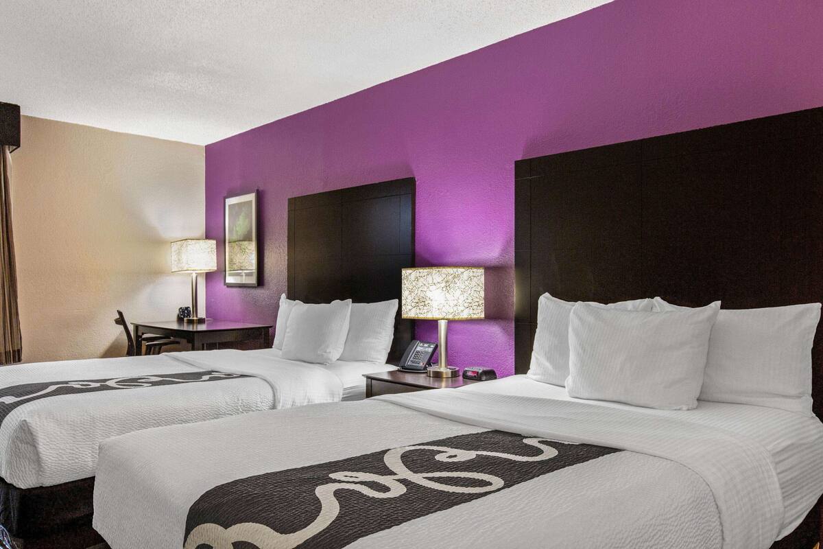 A hotel room with two beds, purple walls, and a nightstand with a lamp.