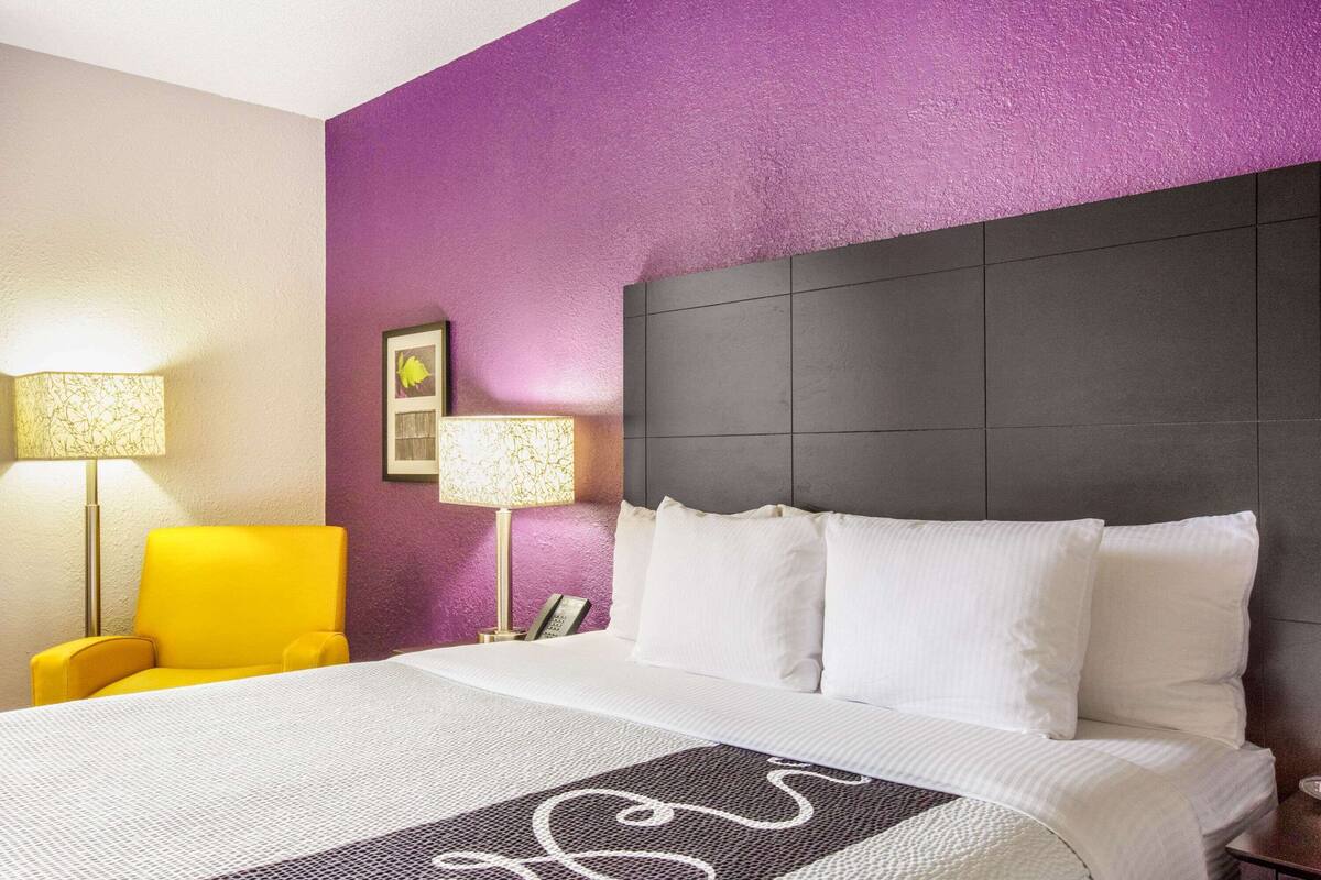 A hotel room with a bed, a yellow armchair, two bedside lamps, and a purple accent wall.