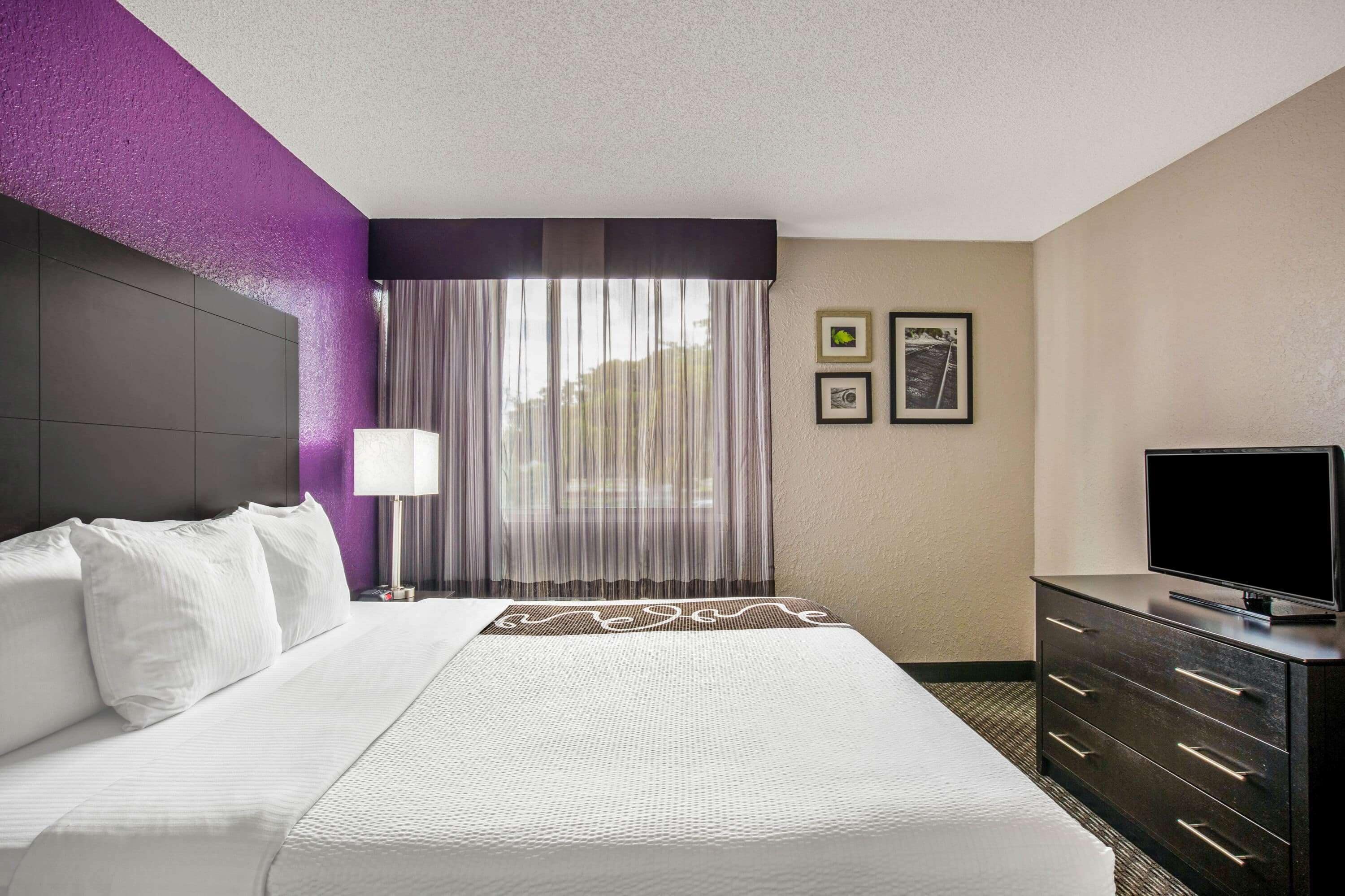 A hotel room with a bed, a TV on a dresser, and a window with curtains.