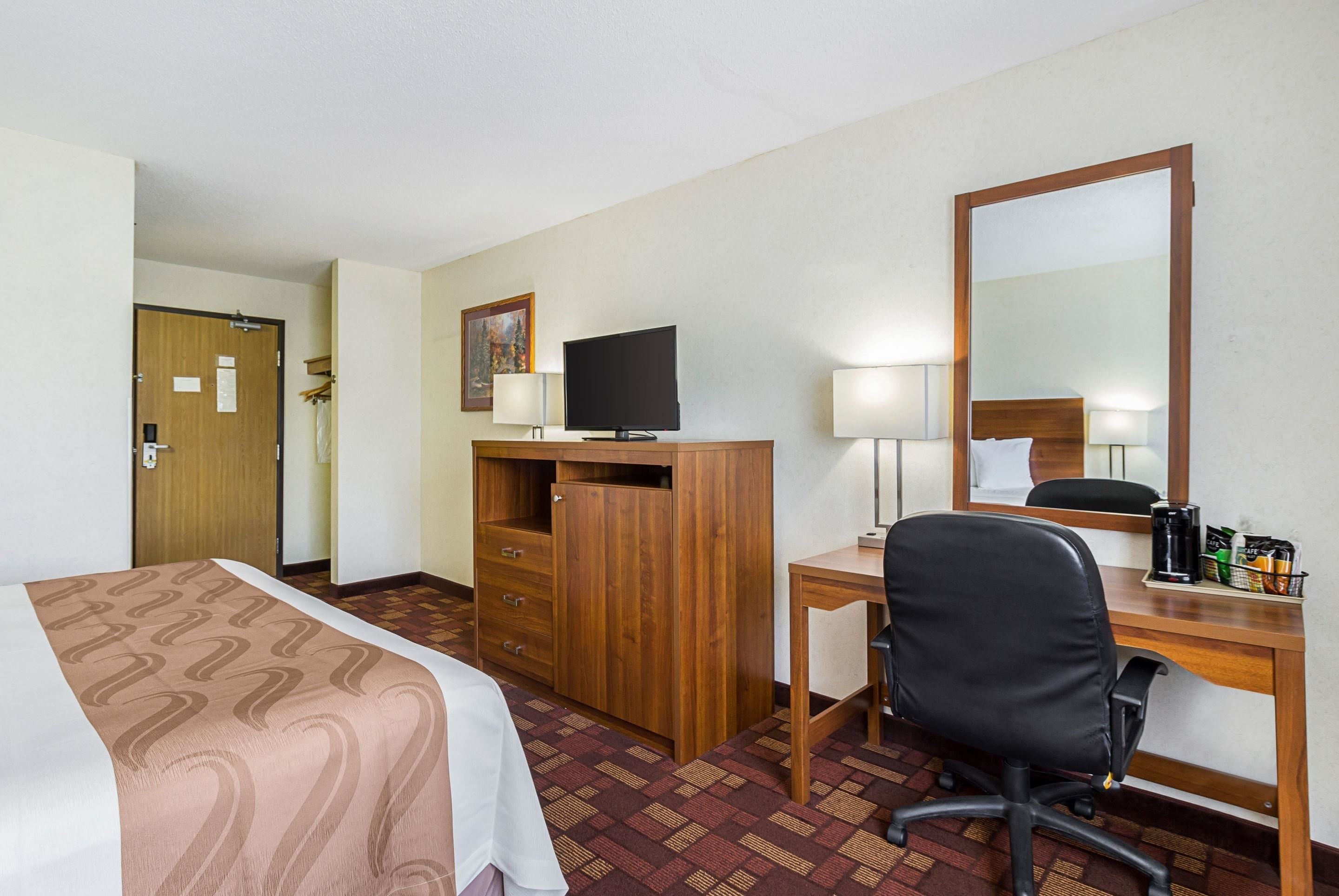 A hotel room with a bed, a desk with a computer, a chair, a TV, and a mirror.