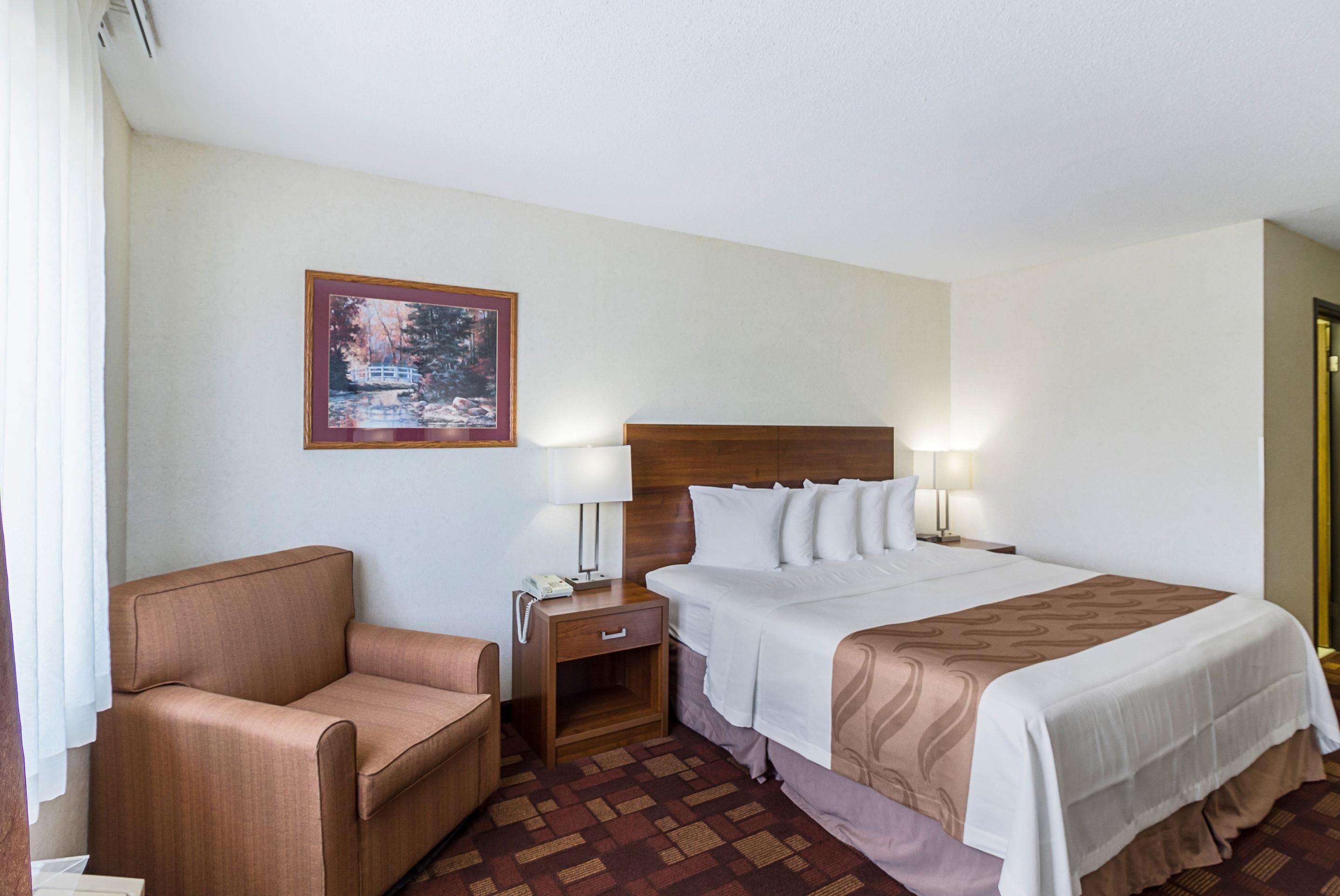 Habitación de hotel con cama, una silla, mesita de noche, lámpara y un cuadro en la pared.