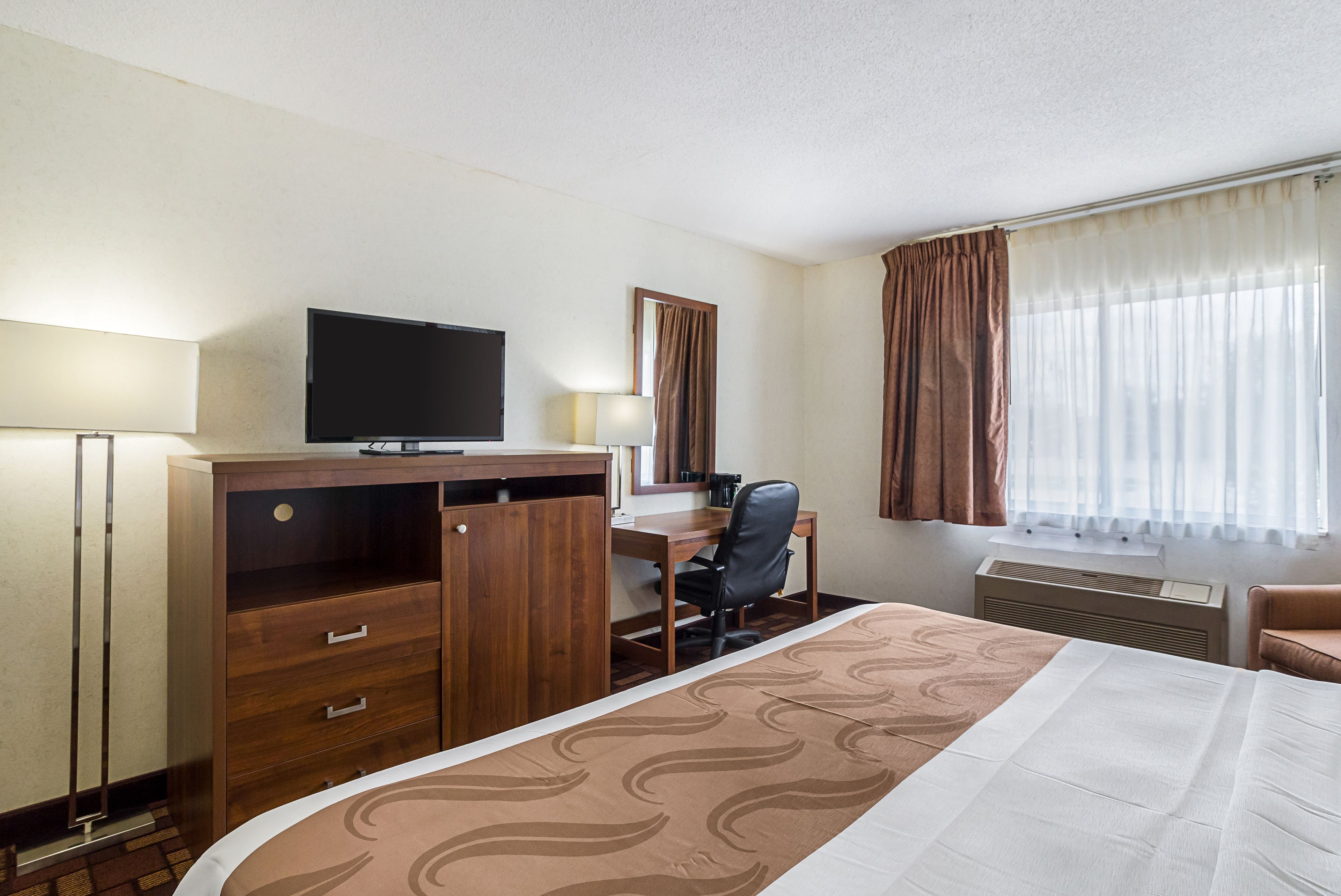 Une chambre d’hôtel comprenant un lit, un bureau avec une chaise, une télévision, un miroir et une fenêtre avec des rideaux.