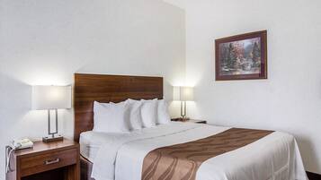 Una cama bien hecha con cabecera de madera, dos lámparas de noche, una mesita con un teléfono y un cuadro en la pared.