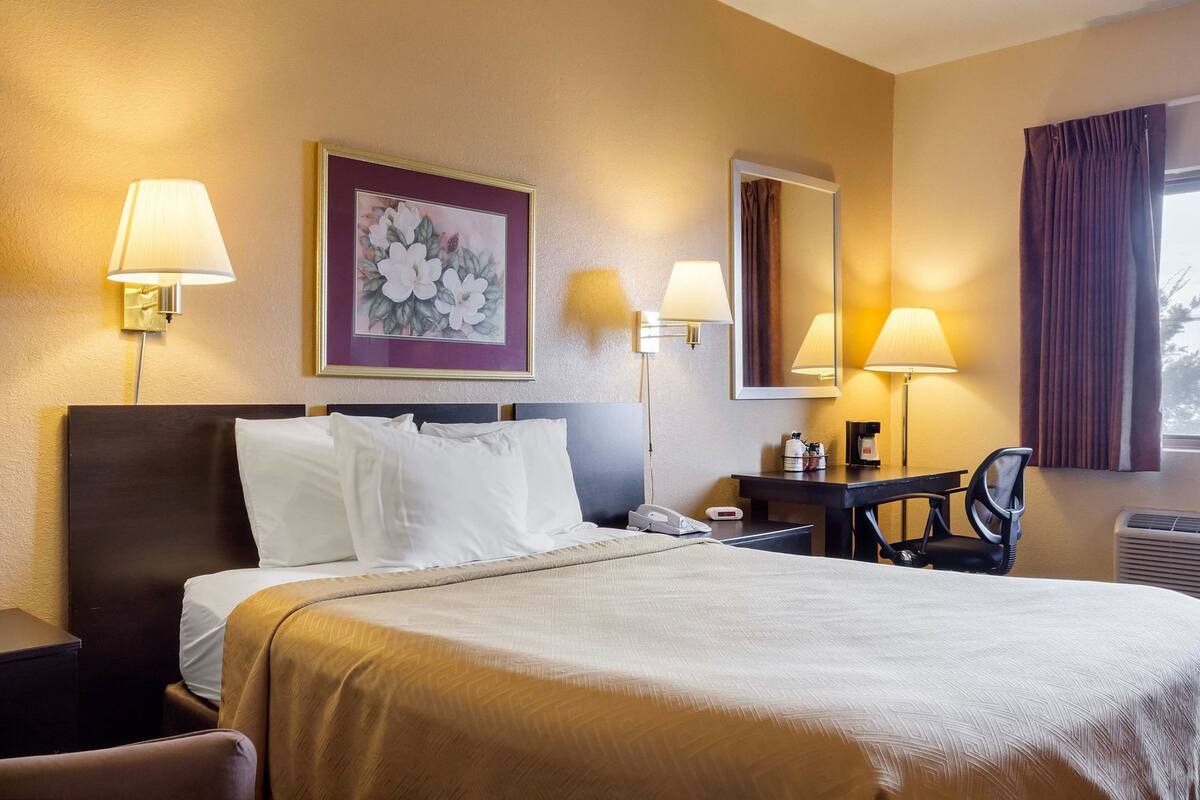 A hotel room with a bed, two bedside lamps, a desk with a chair, a mirror, and a framed floral artwork on the wall.