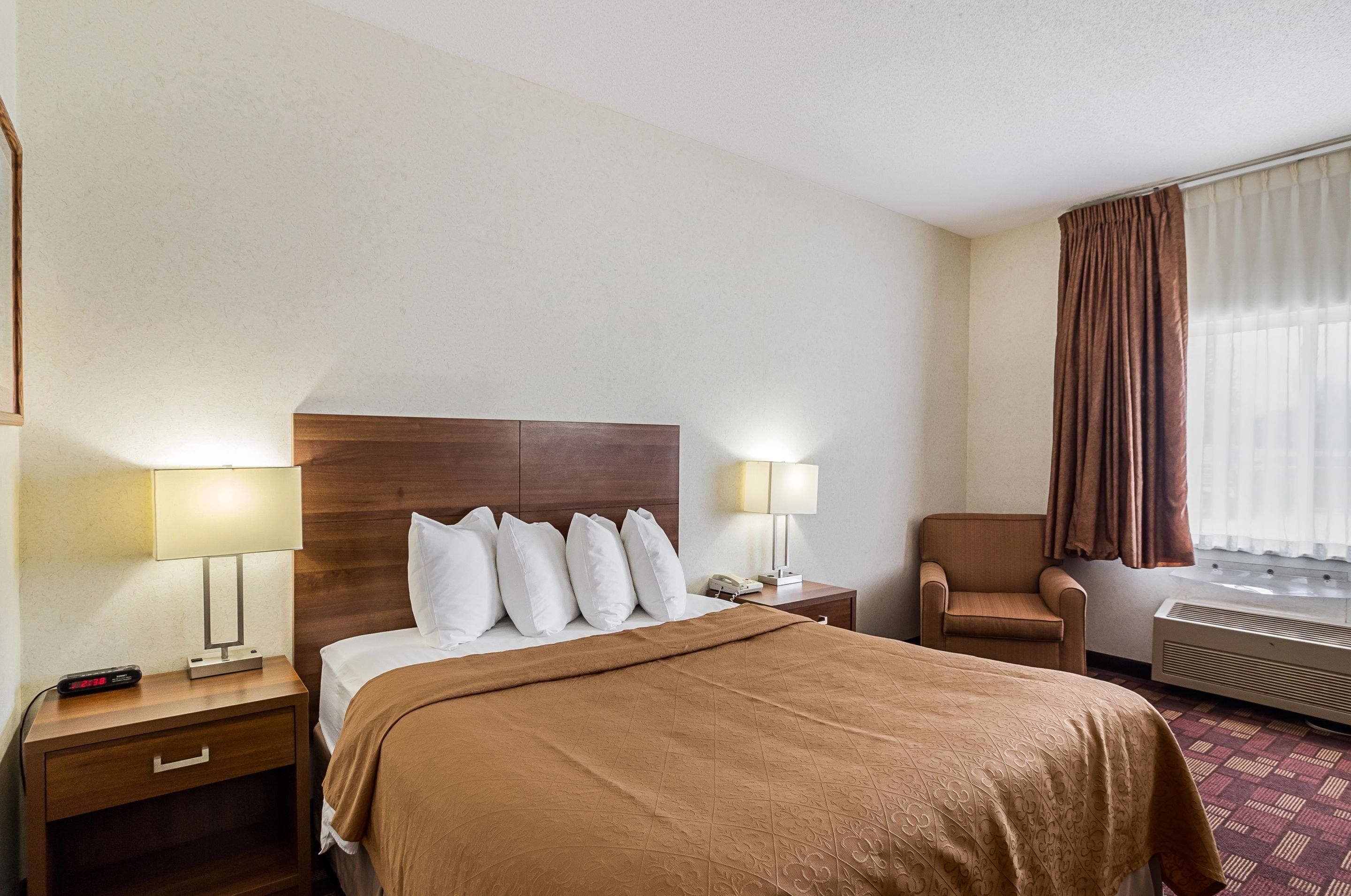 Una habitación de hotel con una cama, dos mesitas de noche con lámparas, una silla, un equipo de aire acondicionado y una ventana con cortinas.