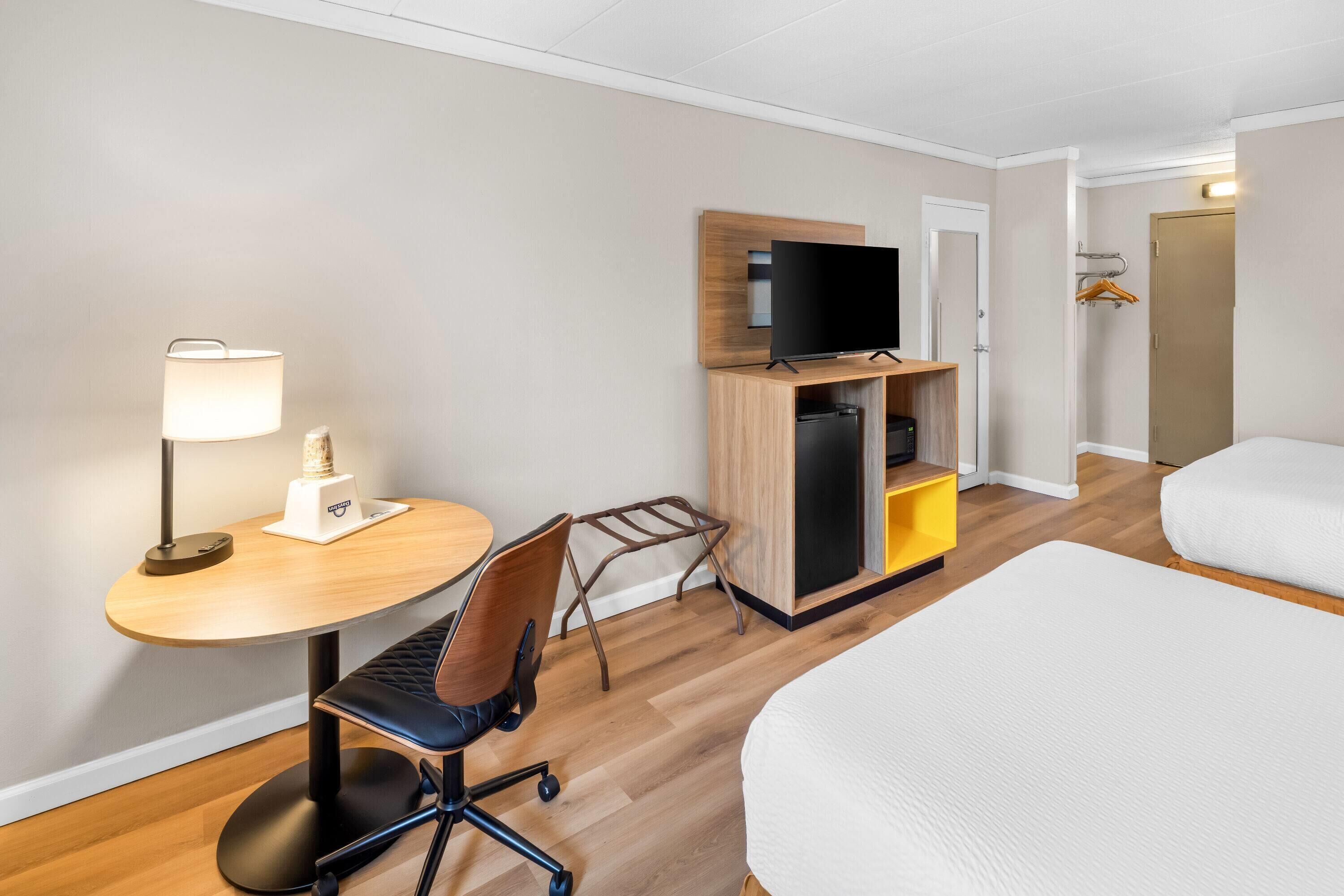 A hotel room with a bed, a desk with a lamp, a chair, a television on a wooden stand, and a wardrobe.