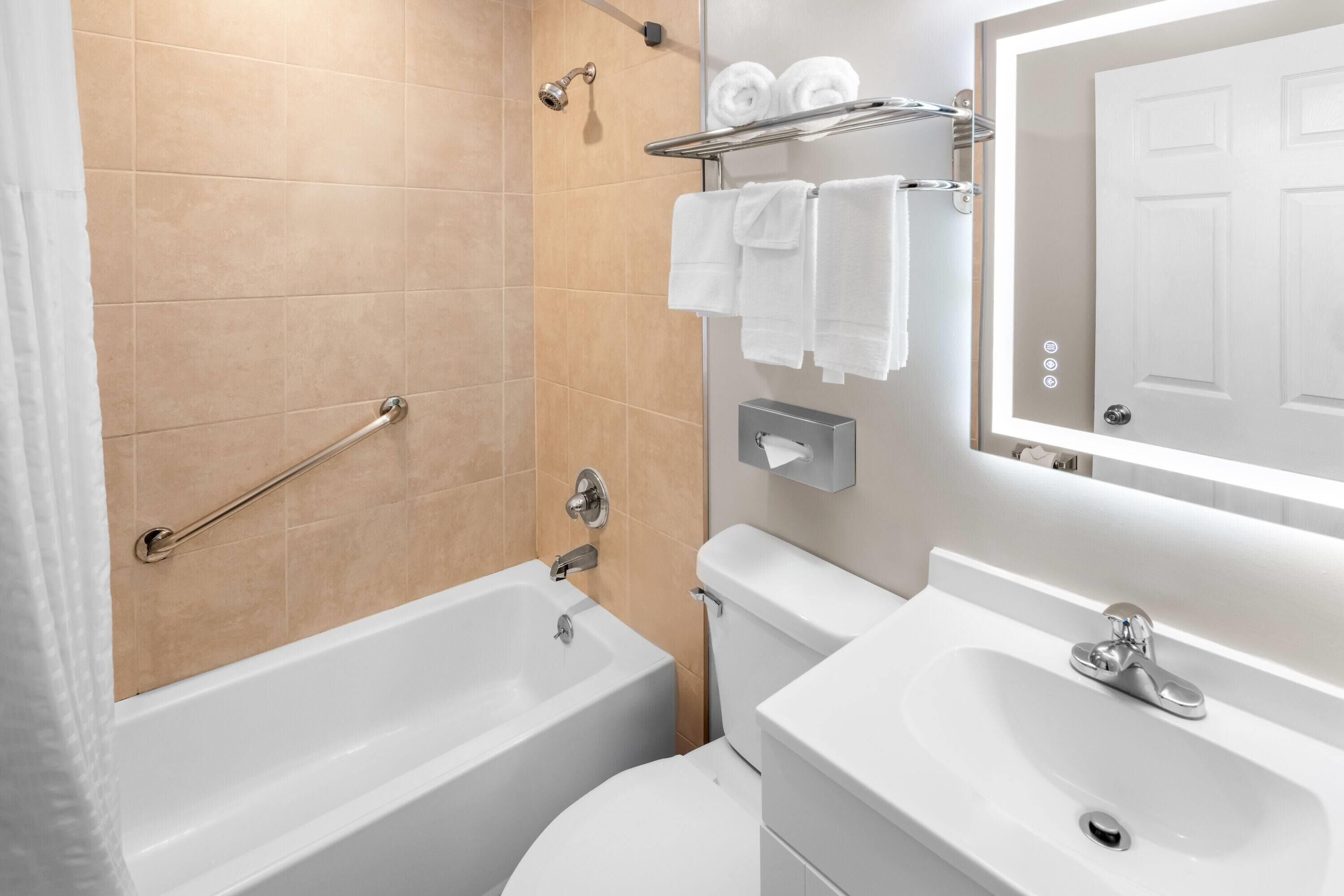 Un baño moderno con bañera, inodoro y lavabo. Hay un perchero con toallas, un cabezal de ducha y un dispensador de papel higiénico.