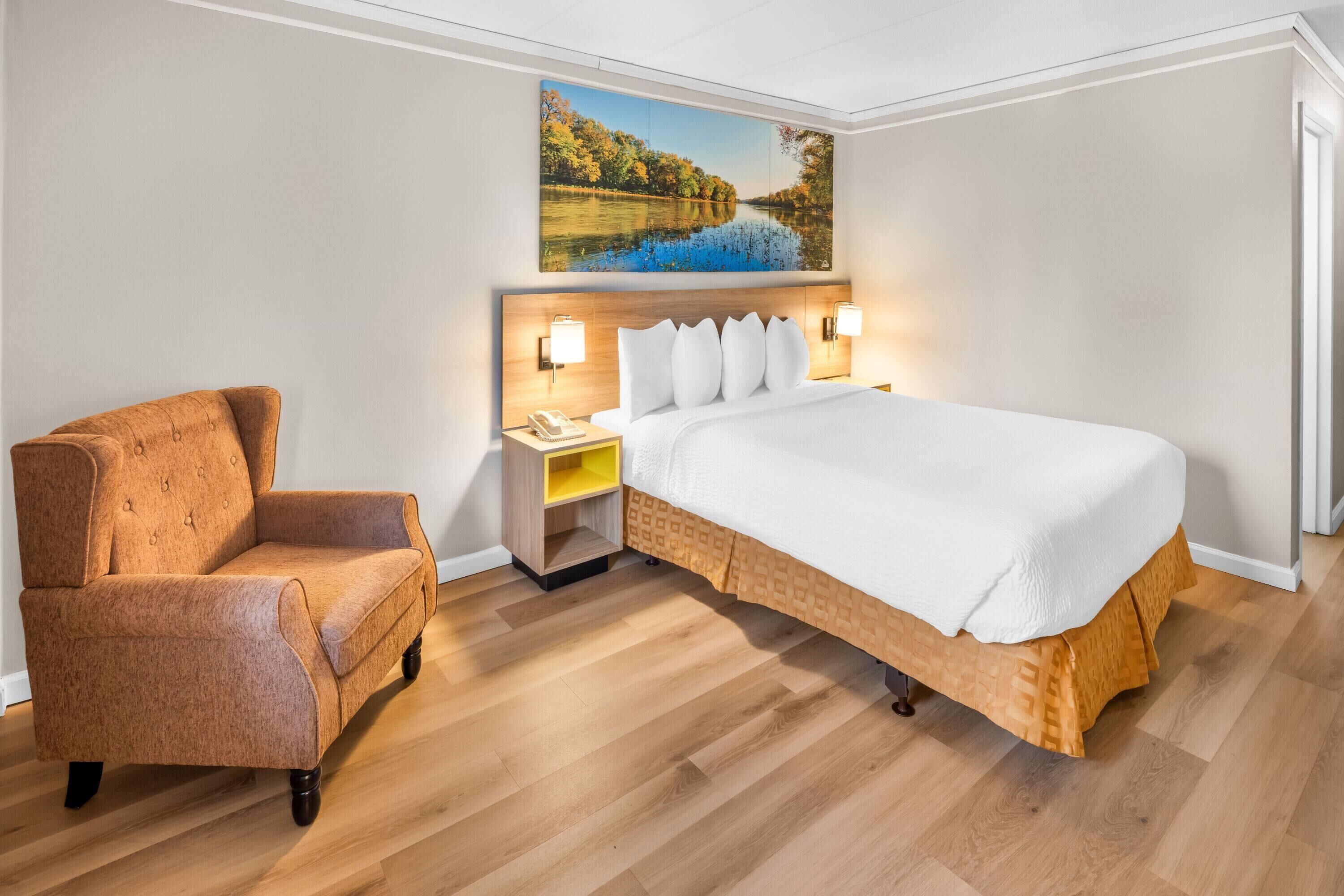 A hotel room with a bed, a chair, a nightstand, and a painting on the wall.