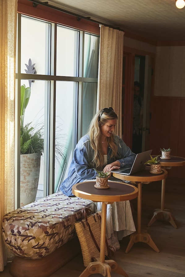 A person sitting on a cushioned bench at a wooden table with a laptop, near a window with curtains, in a room with wooden flooring and a large potted plant.