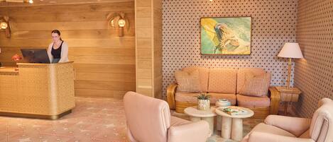 Un área de recepción con un escritorio de madera, una recepcionista, un sofá, dos sillones y una mesita con una lámpara.