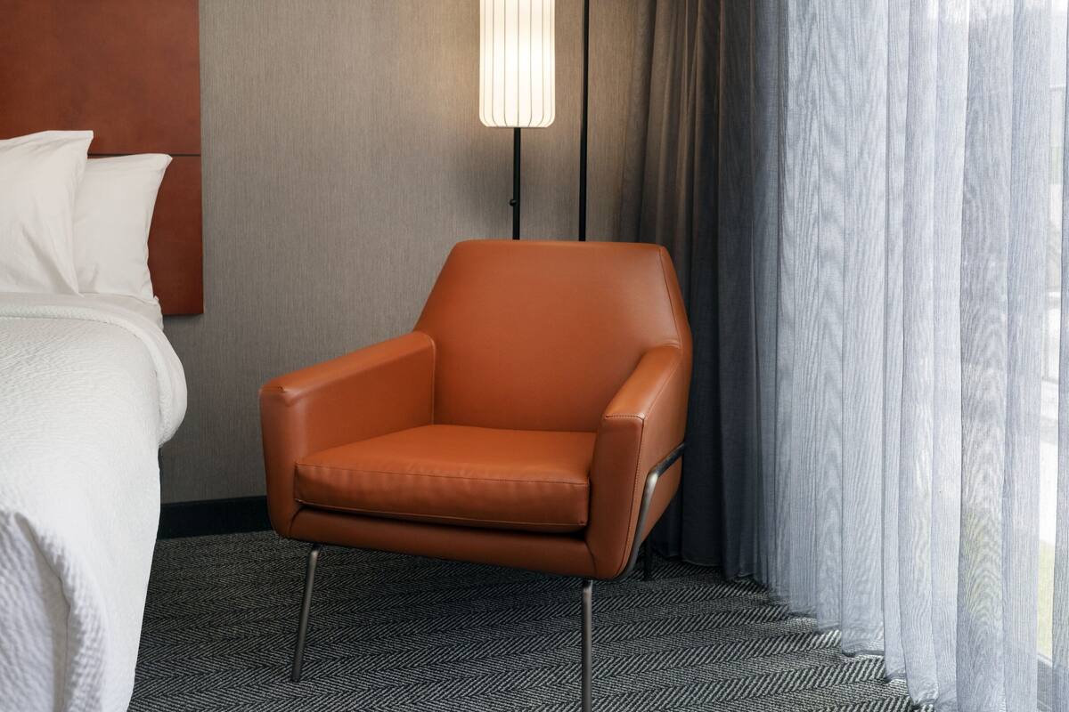 Ein Hotelzimmer mit braunem Ledersessel, einem weißen Bett, einem grauen Teppich und einer modernen Lampe.