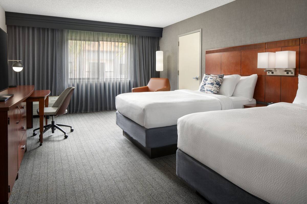 Ein Hotelzimmer mit zwei Betten, einem Schreibtisch, einem Stuhl und einem großen Fenster mit Vorhängen.