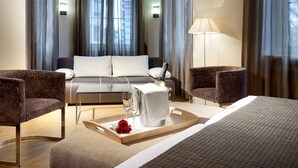 Una habitación de hotel moderna con cama, dos sillones, un sofá, una mesita de noche con lámpara, una bandeja con vaso de agua y una rosa, y una ventana con cortinas.