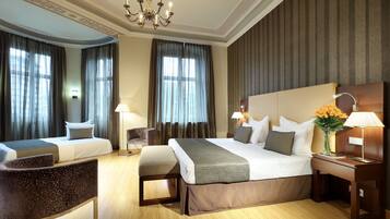 A hotel room with two beds, a desk, a chair, a chandelier, and a nightstand with a lamp and flowers.