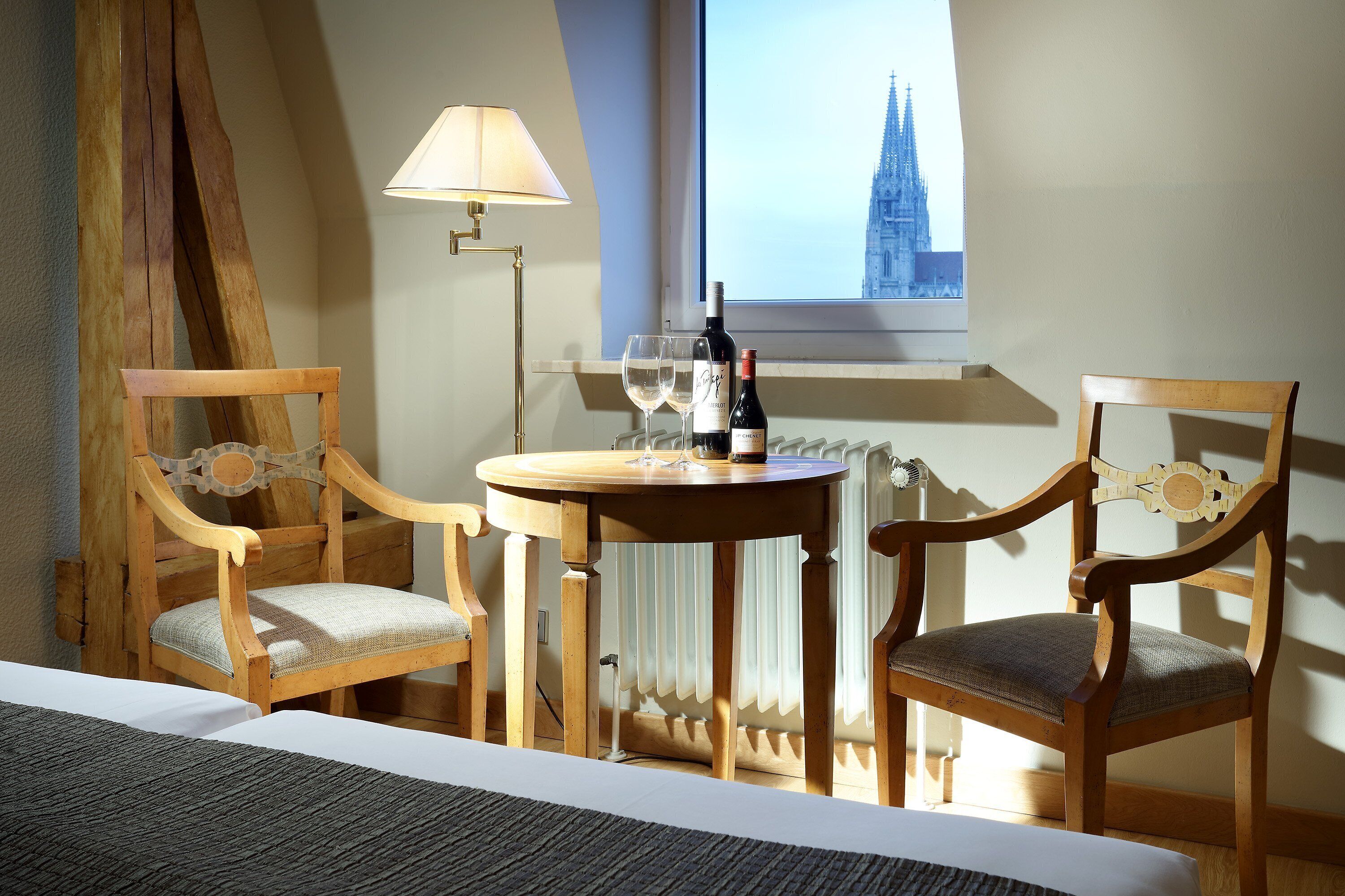 A hotel room with two wooden chairs, a small round table with a bottle of wine and two glasses, a lamp, and a view of a cathedral through a window.