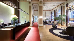 Vestíbulo de hotel con mostrador de recepción, alfombra roja, zona de estar y ventanas amplias.