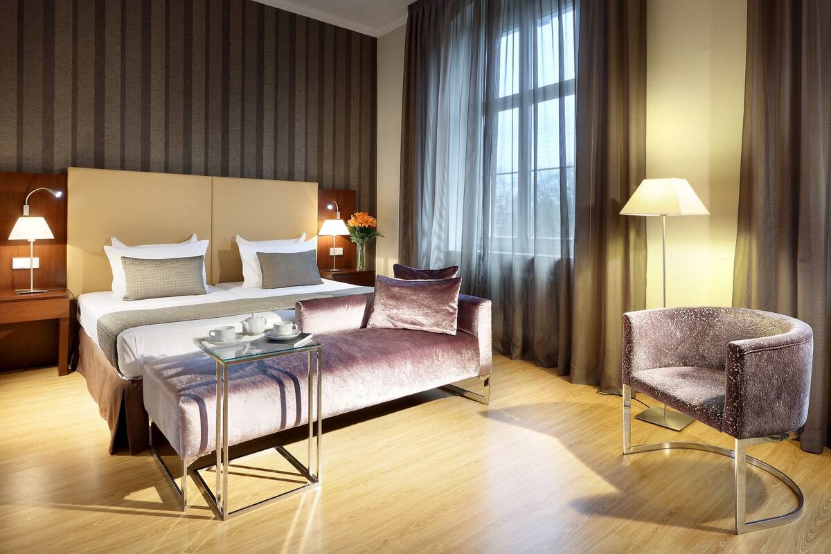 Habitación de hotel con cama, sofá, sillón, mesita de noche con lámparas y ventana con cortinas.