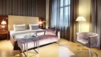 A hotel room with a bed, a sofa, a chair, a bedside table with lamps, and a window with curtains.