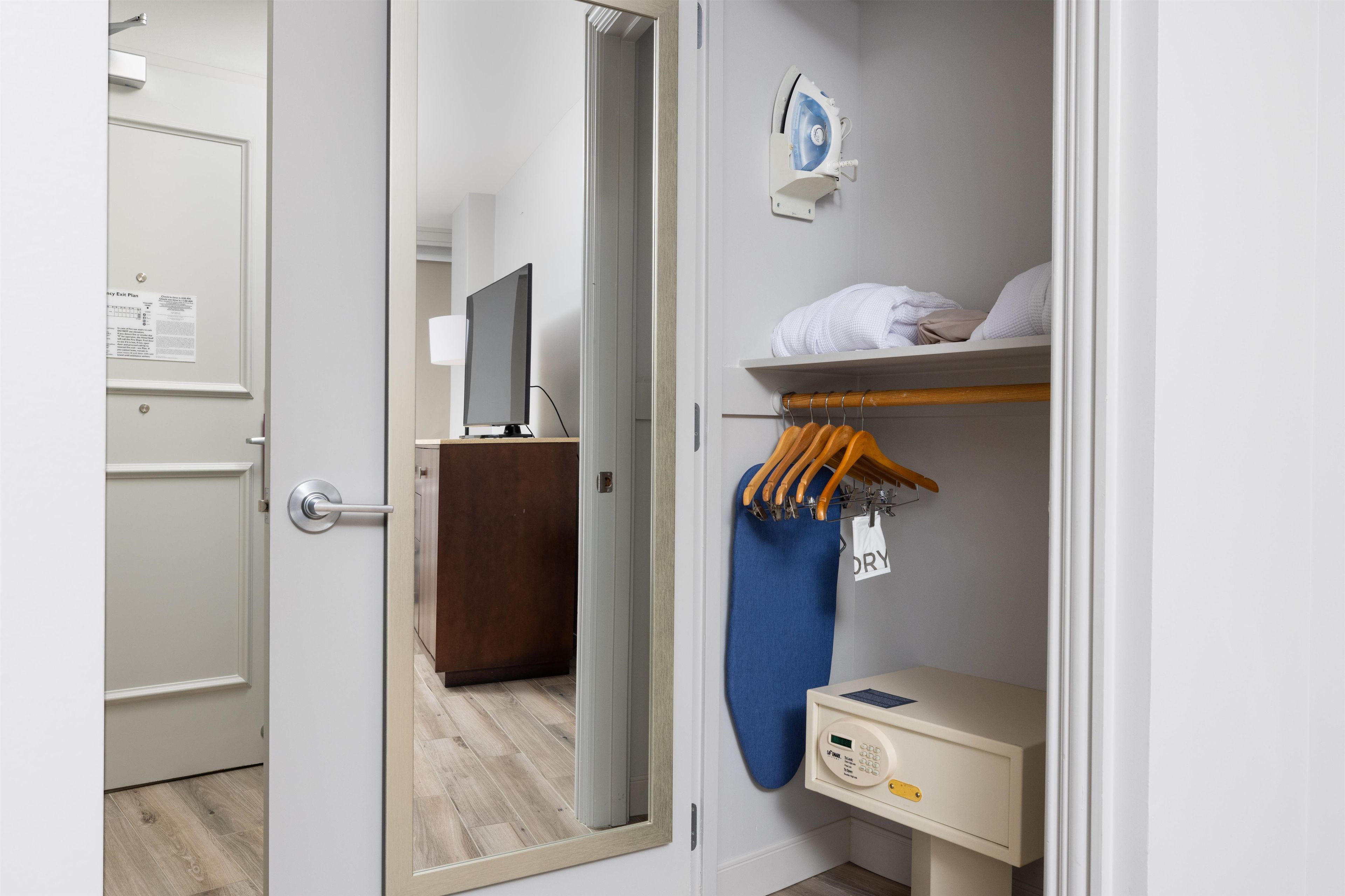 A hotel room with a wardrobe, a mirror, a television, a shelf with towels, and a drying rack.