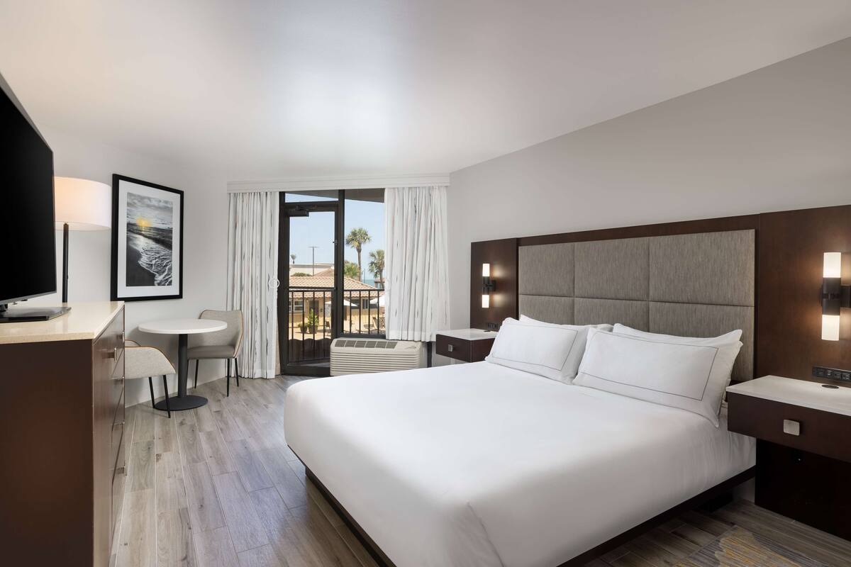 Hotelkamer met een groot bed, een bureau met stoel, een televisie, een ingelijst schilderij en een balkon met gordijnen.