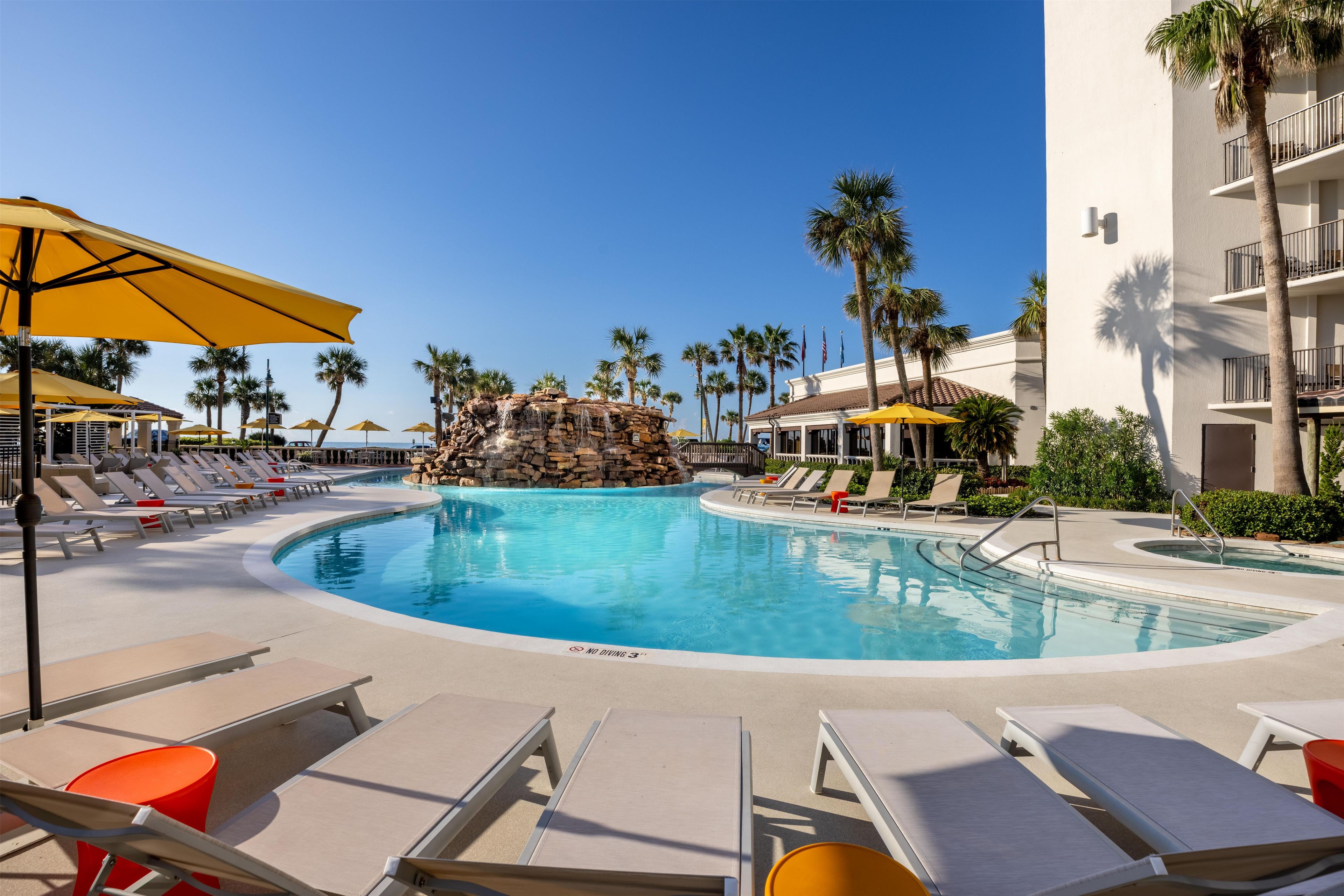 A pool area with lounge chairs and umbrellas, a rock waterfall feature, and palm trees.