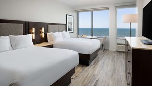 A hotel room with two beds, a desk, a chair, and a large window with a view of the sea.