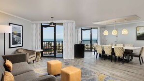 A modern hotel room with a balcony, a dining area, and a living room with a sofa and ottomans.