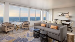 A modern living room with a large window overlooking the sea, a gray sofa, two armchairs, a small round table, and a dining area with a white table and patterned chairs.