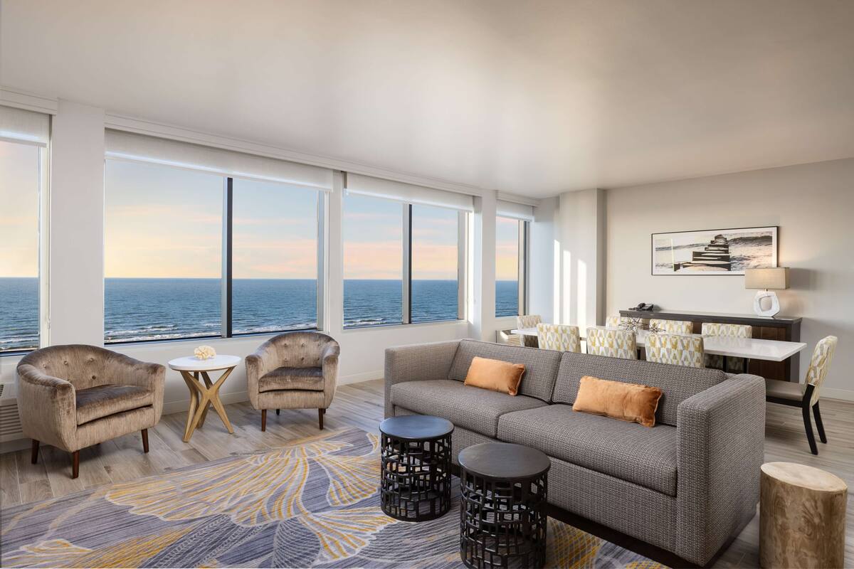 Een moderne woonkamer met een groot raam met uitzicht op zee, een grijze bank, twee fauteuils, een klein rond tafeltje en een eethoek met een witte tafel en gestreepe stoelen.