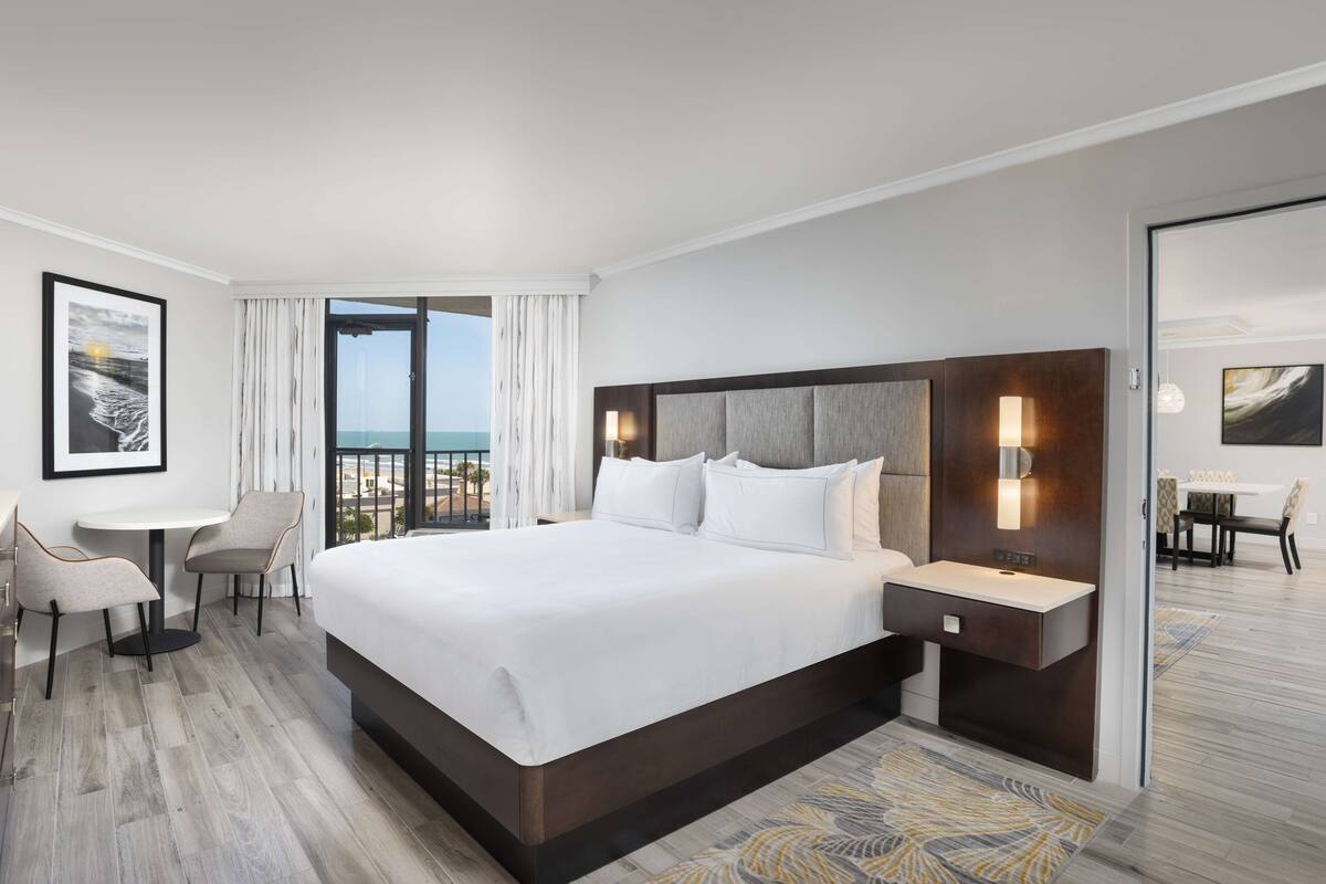 Een moderne hotelkamer met een groot bed, een klein bureau, twee stoelen, uitzicht op de oceaan en een eetgedeelte zichtbaar door een geopende deur.