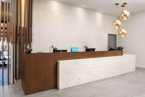 Un comptoir d’accueil d’hôtel avec un comptoir en bois et un banc en marbre, une enseigne bleue et trois luminaires sphériques suspendus.