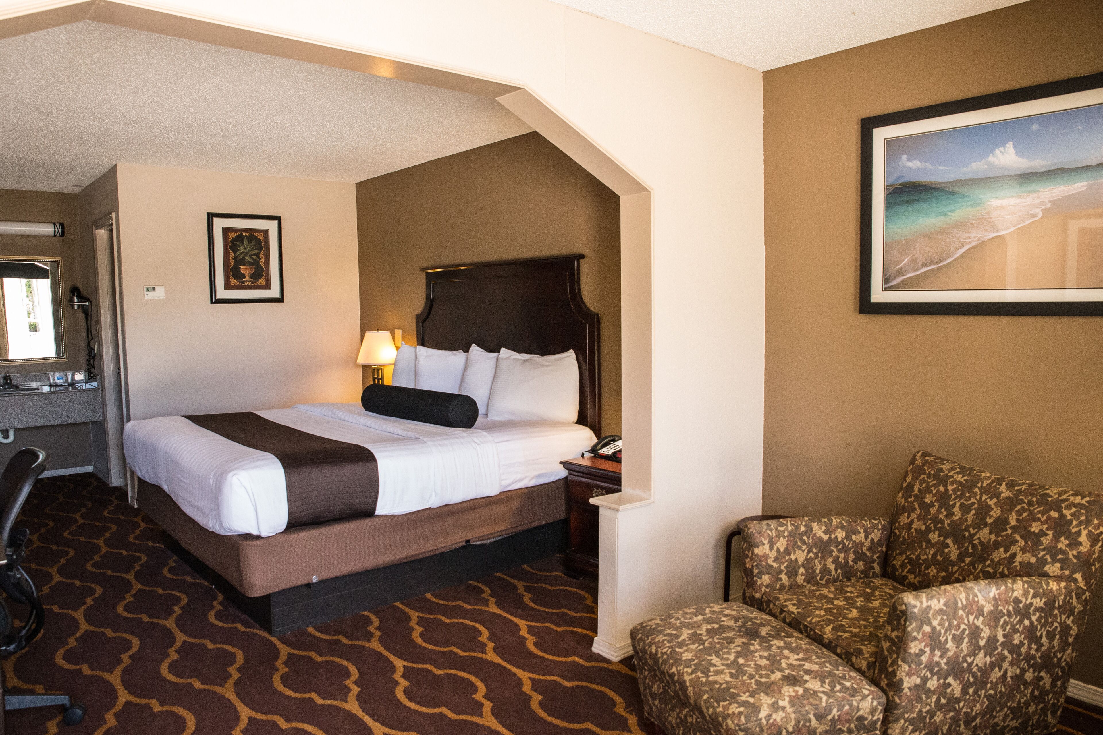 A hotel room with a large bed, a desk with a lamp, a chair, and a framed picture of a beach.