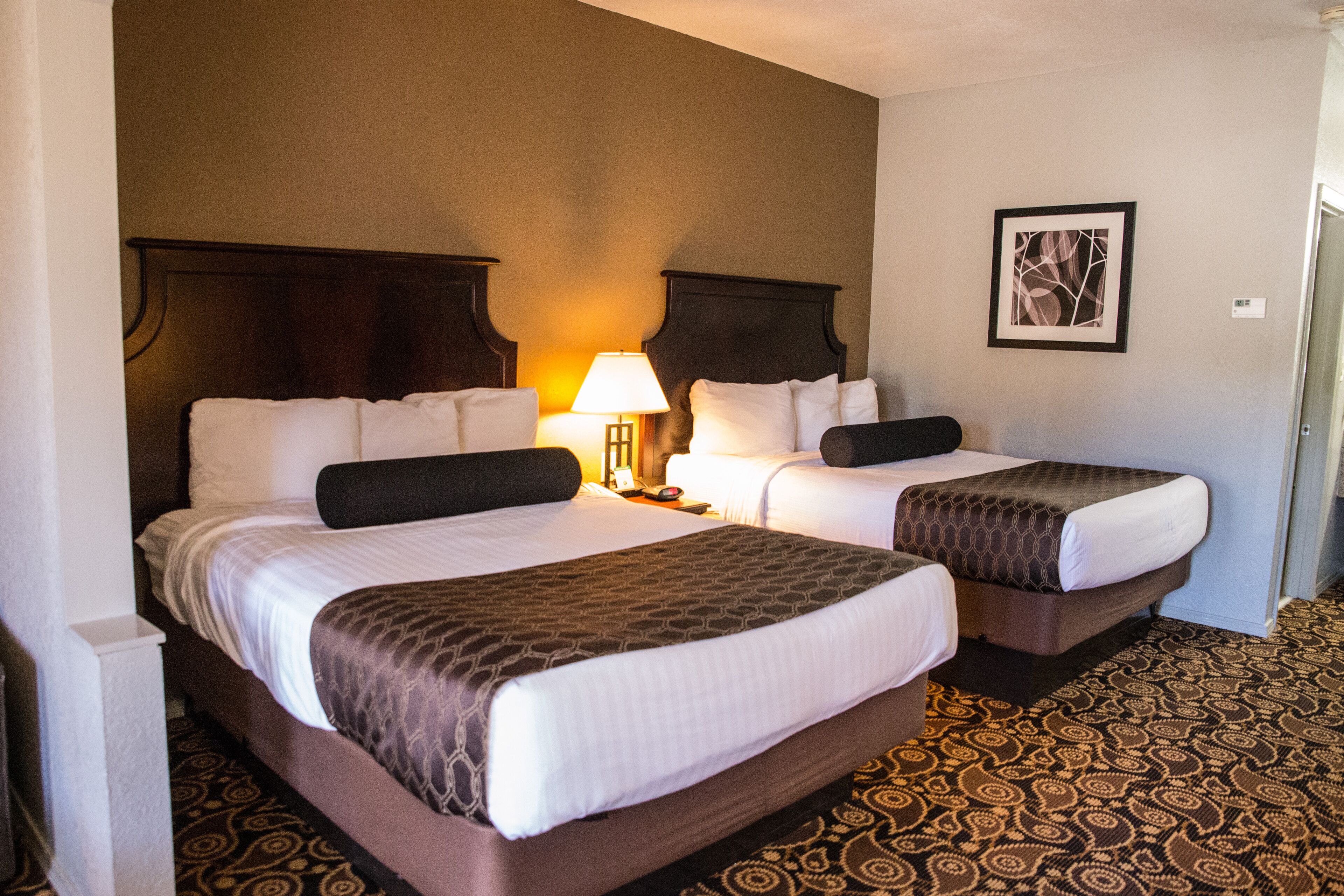 Habitación de hotel con dos camas, una mesita de noche con lámpara, un cuadro enmarcado en la pared y alfombra estampada.