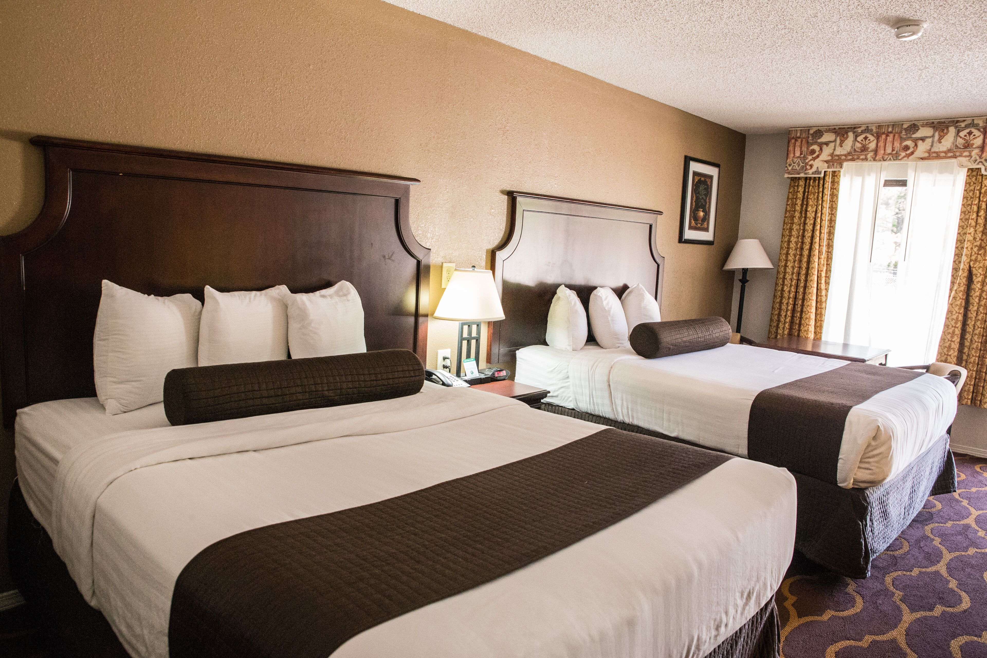 A hotel room with two beds, each with a wooden headboard, white bedding, and a brown accent pillow. There are bedside lamps, a framed picture on the wall, and a window with curtains.