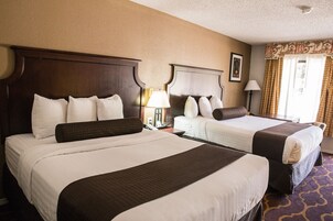 Habitación de hotel con dos camas, cada una con cabecero de madera, ropa de cama blanca y almohada decorativa marrón. Hay lámparas de noche, un cuadro en la pared y una ventana con cortinas.
