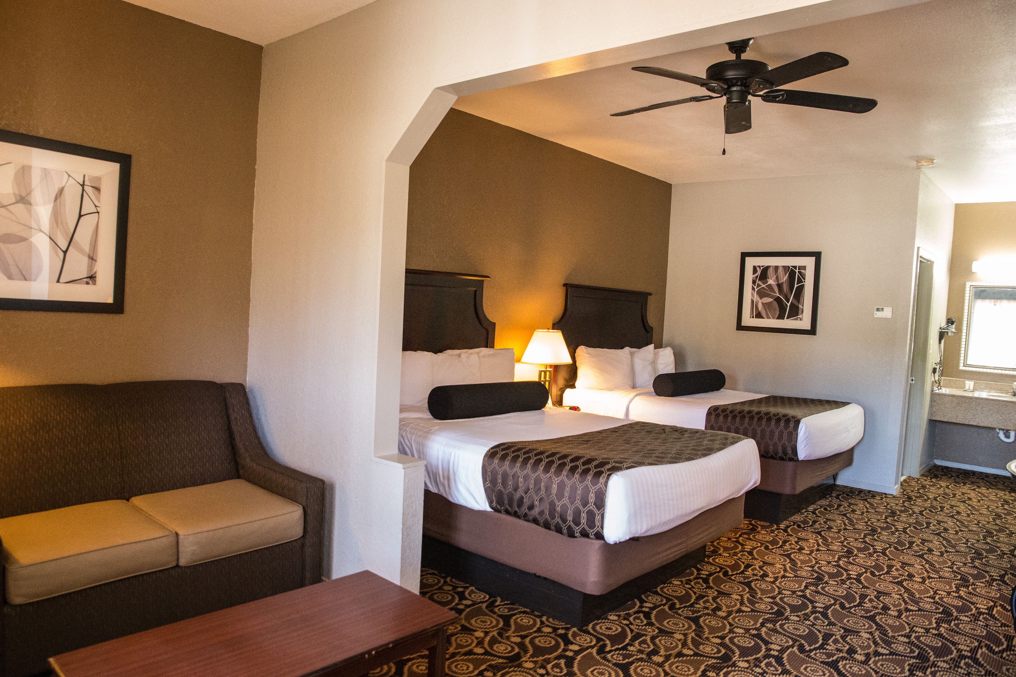 Quarto de hotel com duas camas, um sofá, uma escrivaninha e um ventilador de teto.