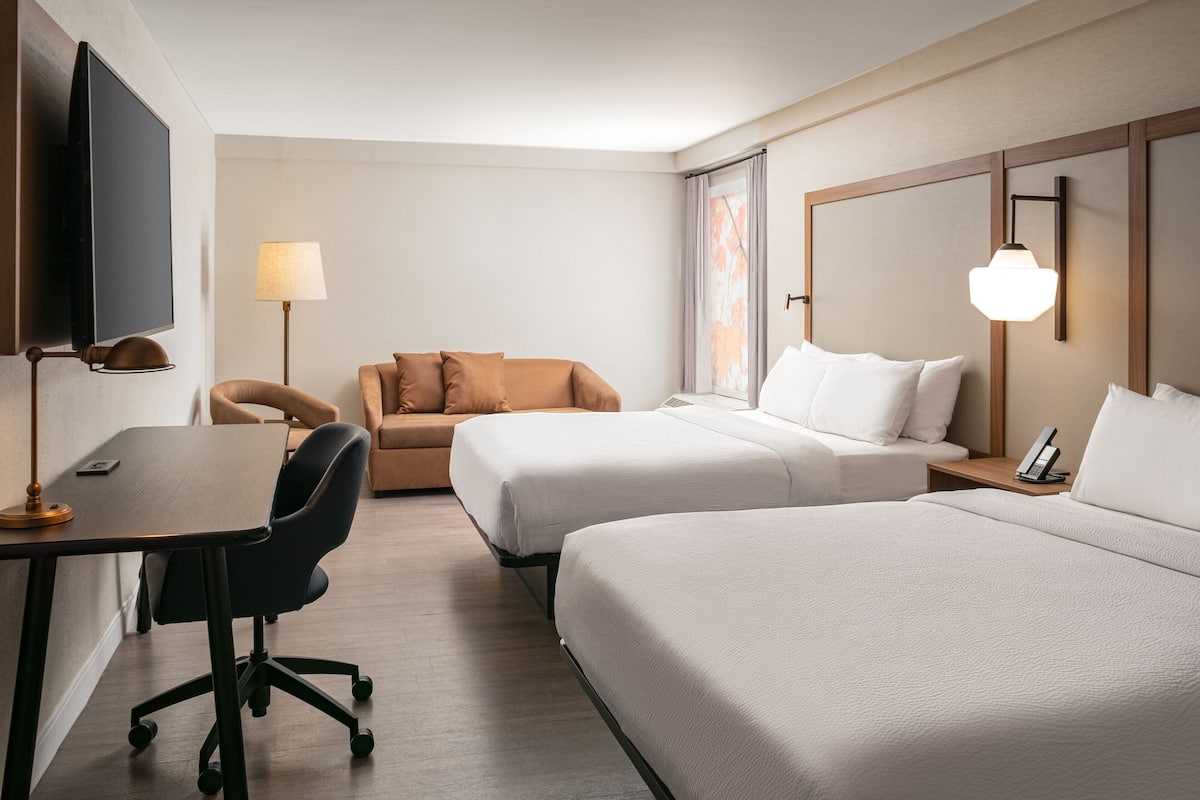 A hotel room with two beds, a desk, a chair, a TV, and a lamp.
