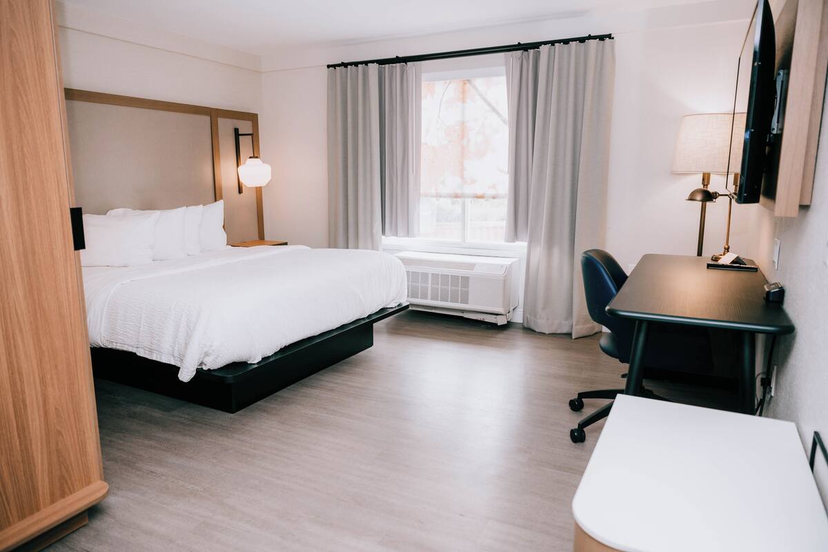 A hotel room with a bed, a desk, a chair, a lamp, and a window with curtains.