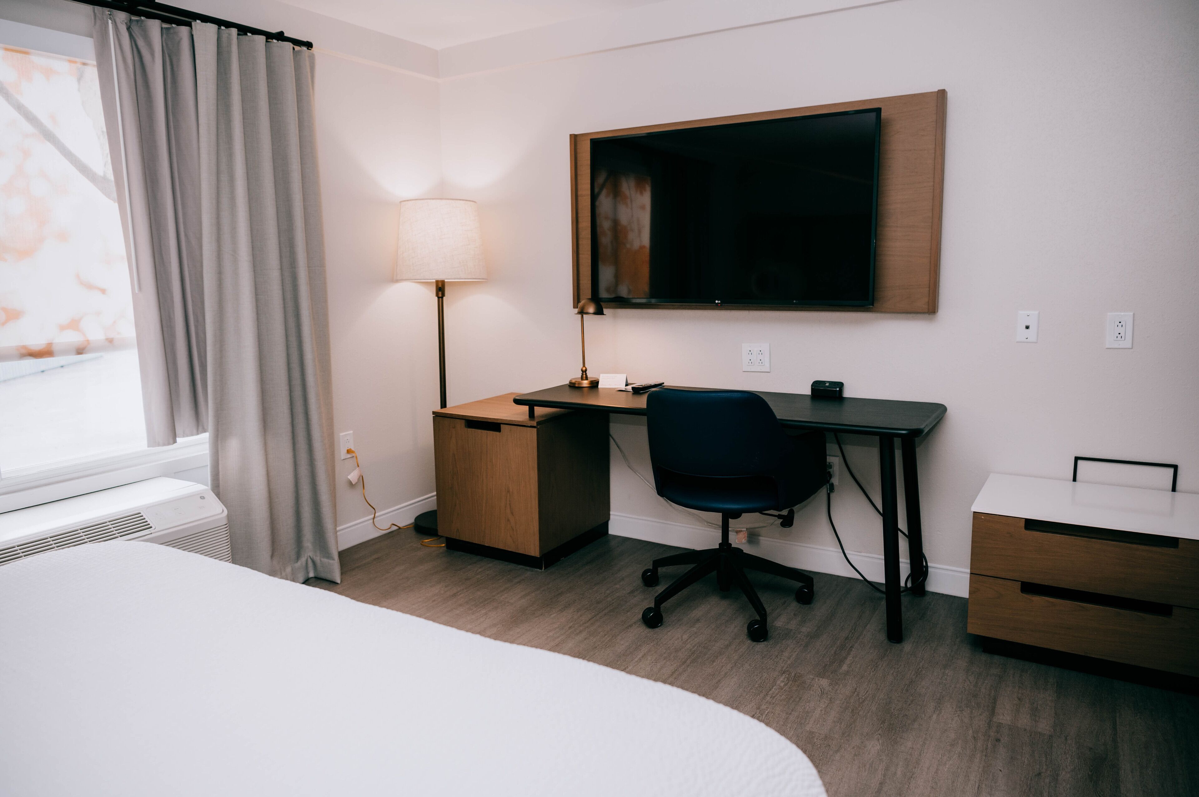 A hotel room with a bed, a desk, a chair, a lamp, and a TV mounted on the wall.