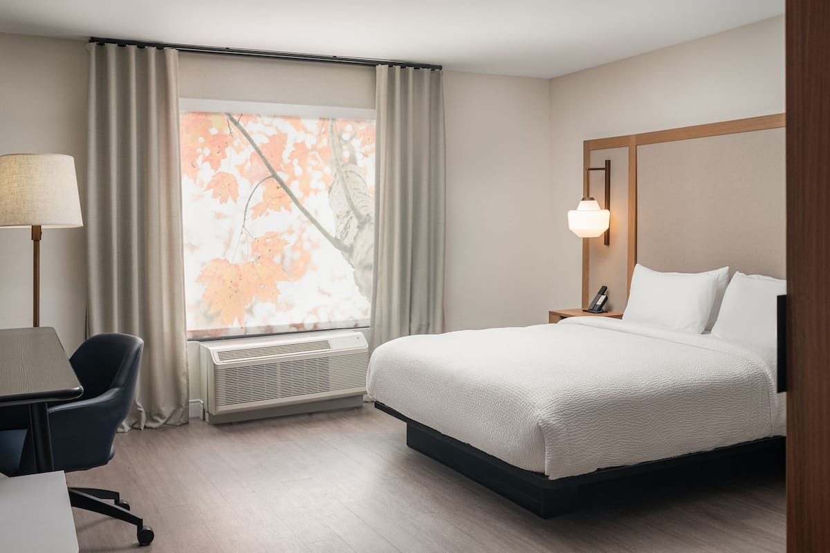 A hotel room with a bed, a desk, a chair, a window with curtains, and a wall-mounted lamp.