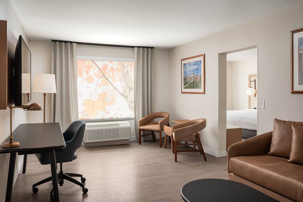 A hotel room with a desk, two chairs, a sofa, a bed, a window with curtains, and framed pictures on the walls.