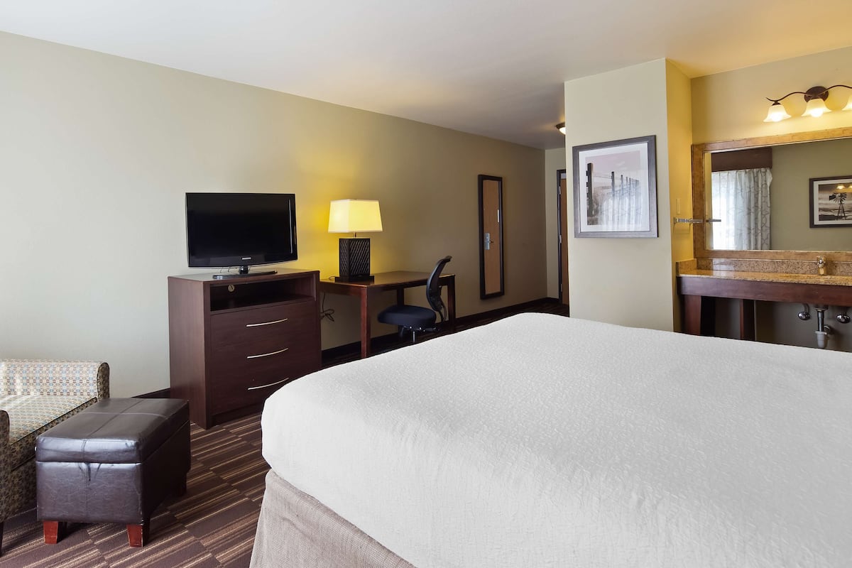 A hotel room with a large bed, a desk with a lamp, a television, a chair, and a bathroom with a mirror and sink.