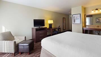 A hotel room with a large bed, a desk with a lamp, a television, a chair, and a bathroom with a mirror and sink.