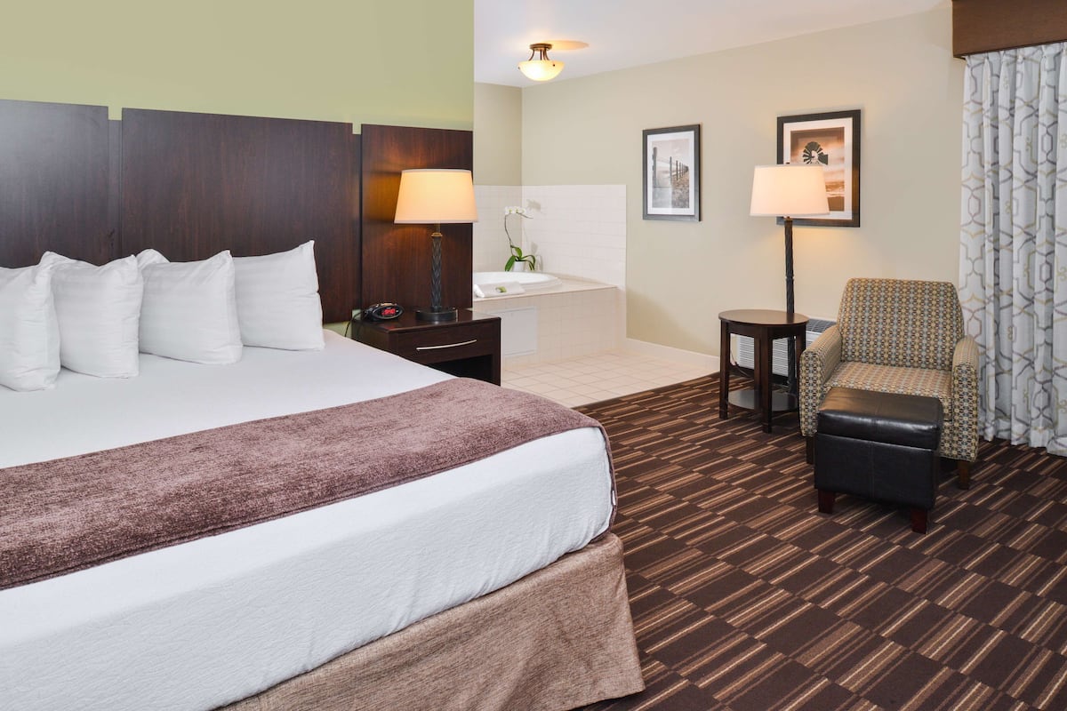 A hotel room with a large bed, a small desk, a chair, a nightstand, a lamp, a picture frame, and a bathroom with a sink and mirror.