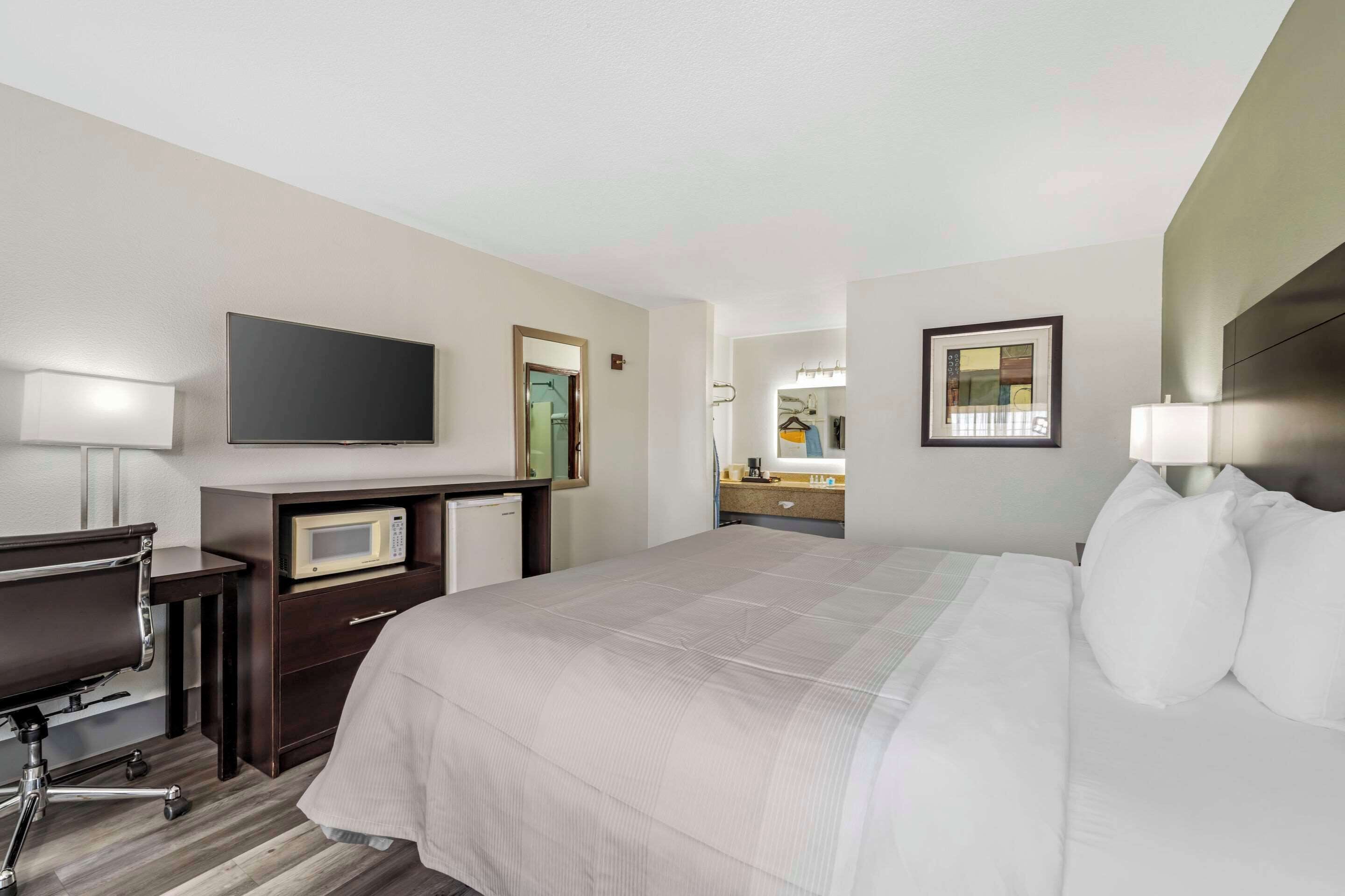 A hotel room with a large bed, a desk with a computer, a microwave, a TV, and a bathroom with a mirror and sink.