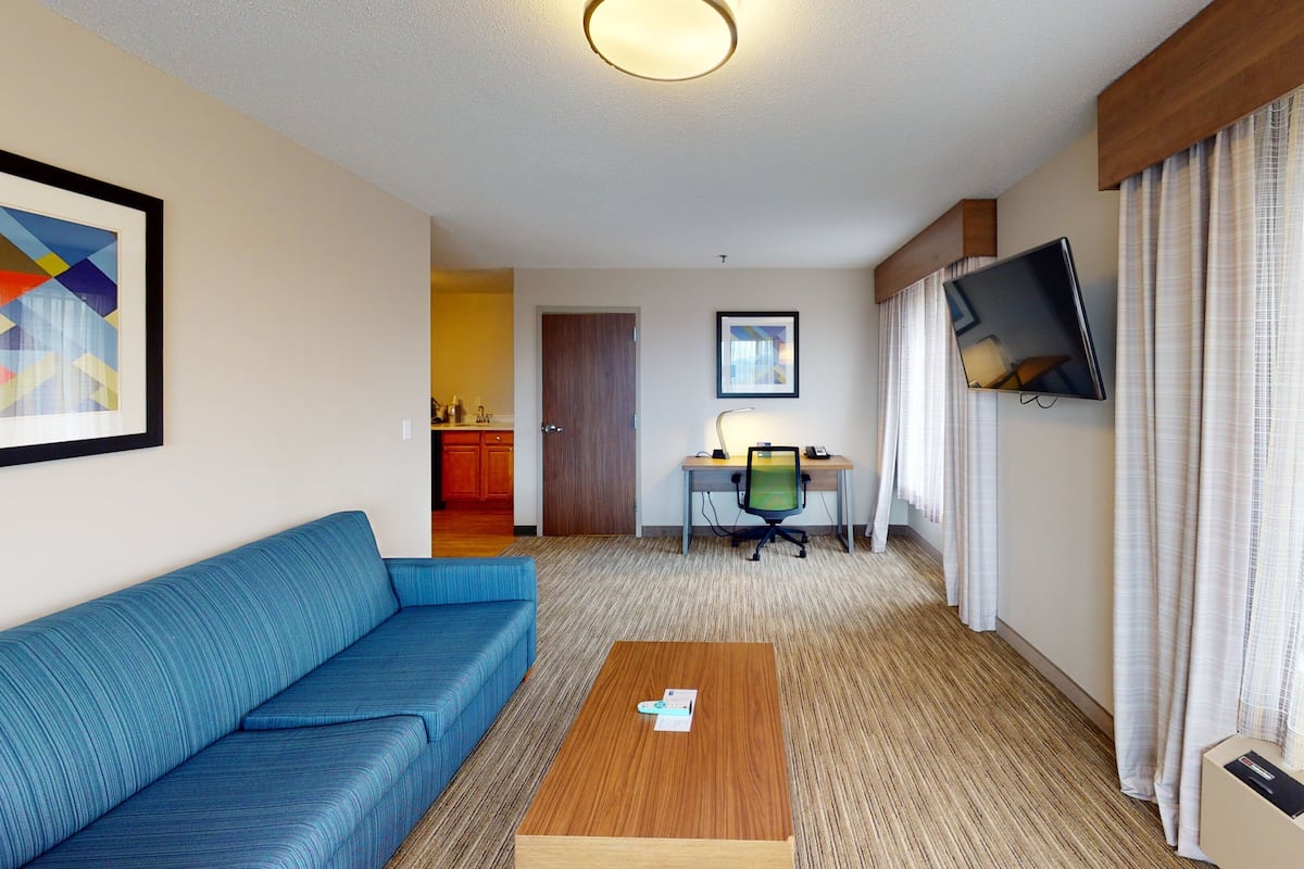 A hotel room with a blue sofa, a wooden coffee table, a desk with a chair, a TV mounted on the wall, and a kitchen area visible through an open door.