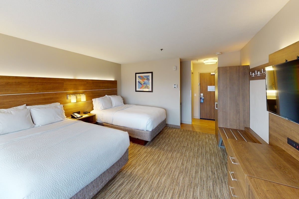 A hotel room with two beds, a wooden headboard, a flat-screen TV, a desk with a lamp, and a door leading to a hallway.