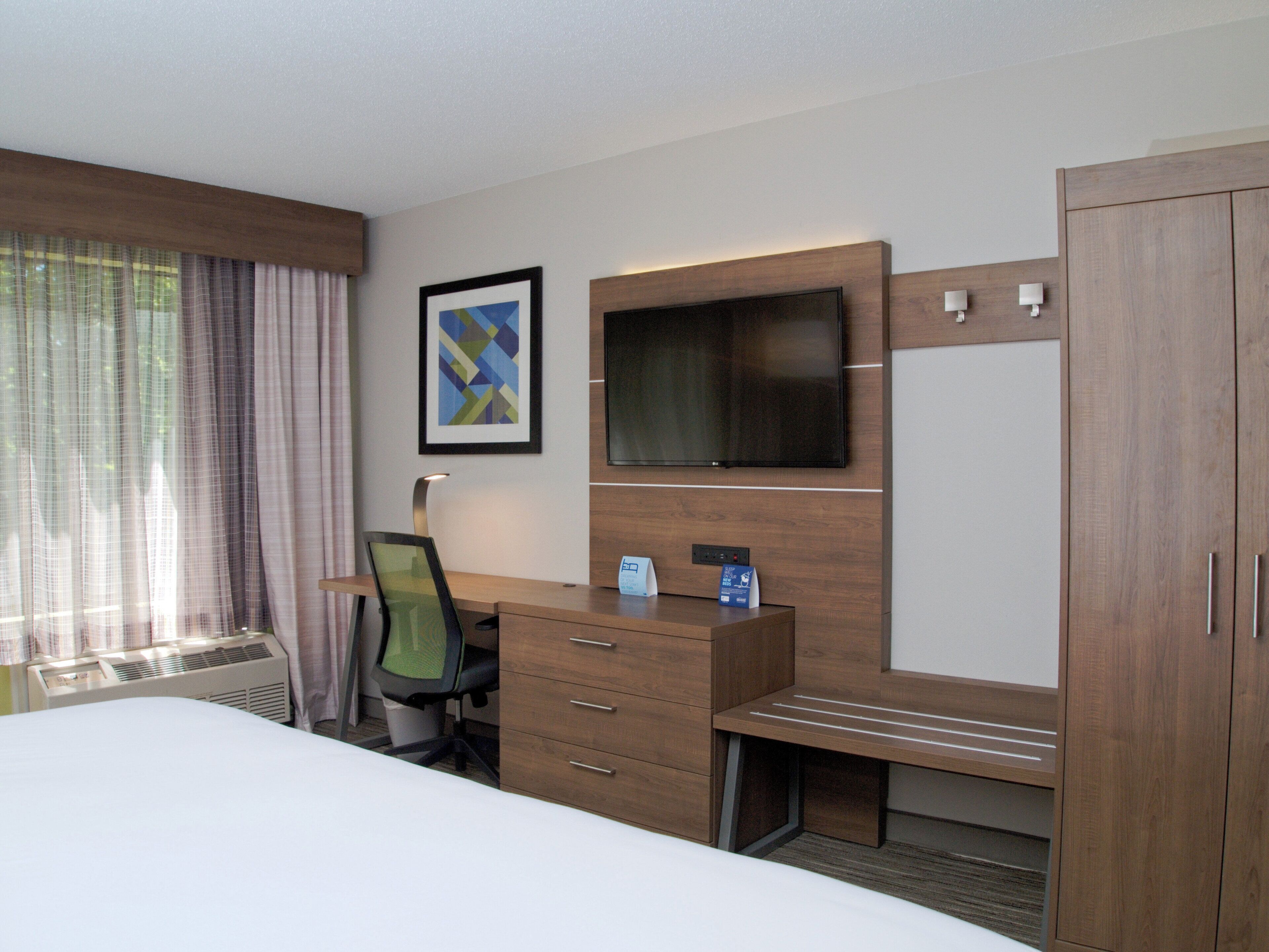 Habitación de hotel con cama, escritorio con cajones, una silla, televisor de pantalla plana, un cuadro enmarco y un armario.