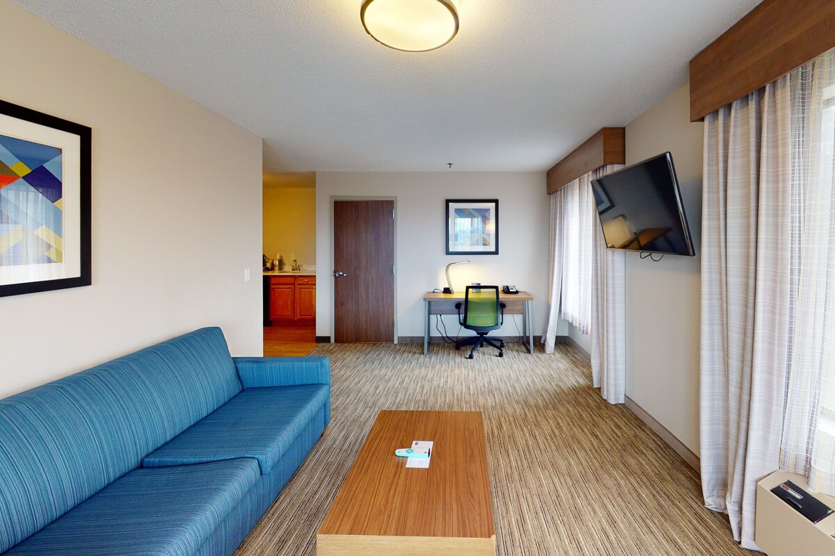 Una camera d'albergo con un divano blu, un tavolino in legno, una scrivania con una sedia, una TV a parete e una zona cucina visibile attraverso una porta aperta.
