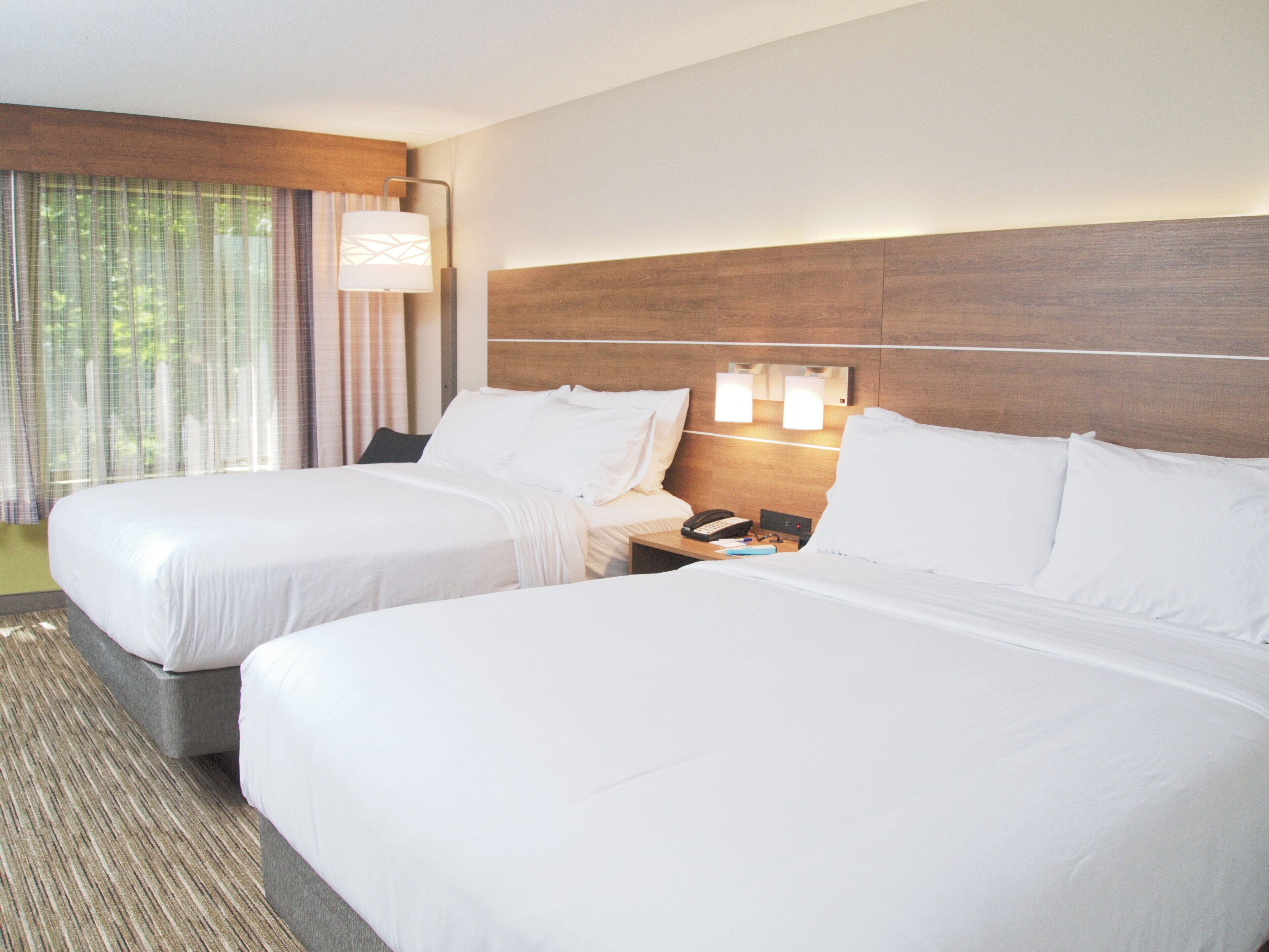 A hotel room with two beds, a nightstand with a phone and lamp, and a window with curtains.